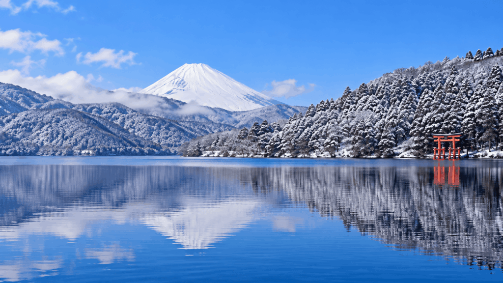 Snow-covered Mount Fuji reflected in Lake Ashi beside Hakone Shrine's red torii gate and snowy forest in winter