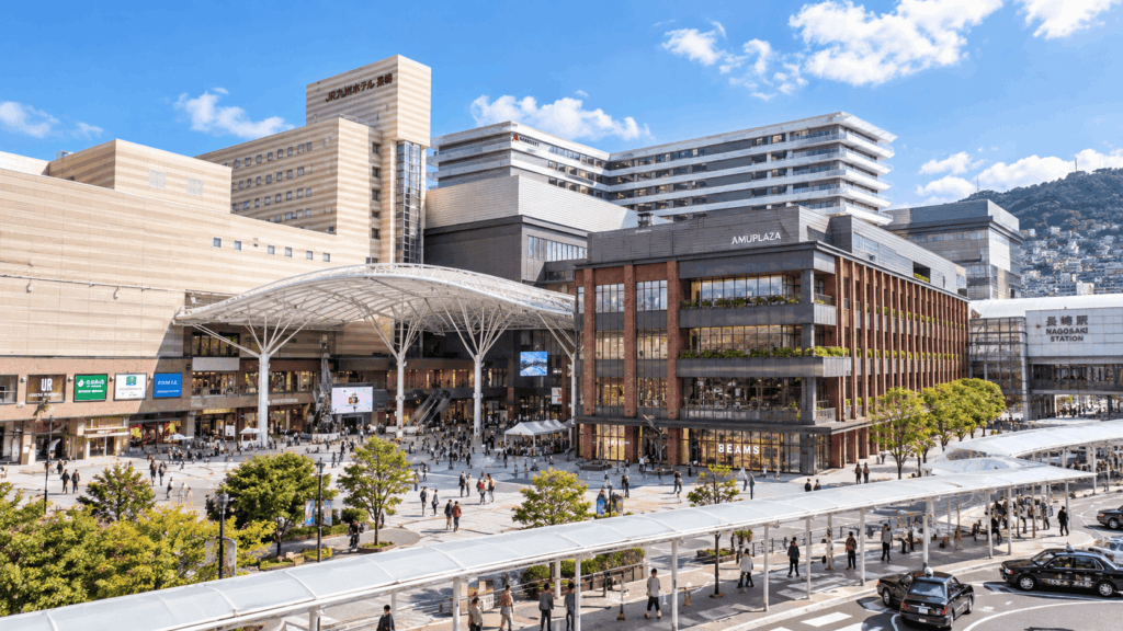 Nagasaki Station and Amu Plaza shopping complex on a sunny day, with modern buildings, a spacious plaza, and pedestrians