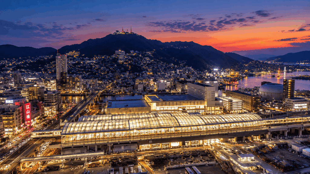 Aerial view of Nagasaki Station illuminated at sunset, with Mount Inasa, city buildings, and Nagasaki Harbor in the background