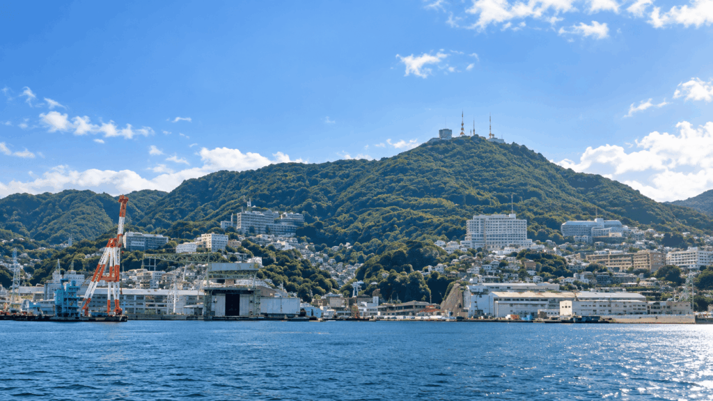 Daytime view of Mount Inasa rising above Nagasaki Harbor, with waterfront buildings, shipyard cranes, and green hills