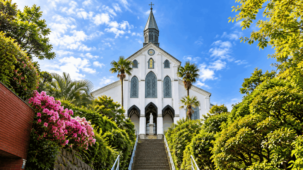 White Gothic-style Oura Cathedral in Nagasaki, framed by lush greenery, pink flowers, palm trees, and a long stone staircase