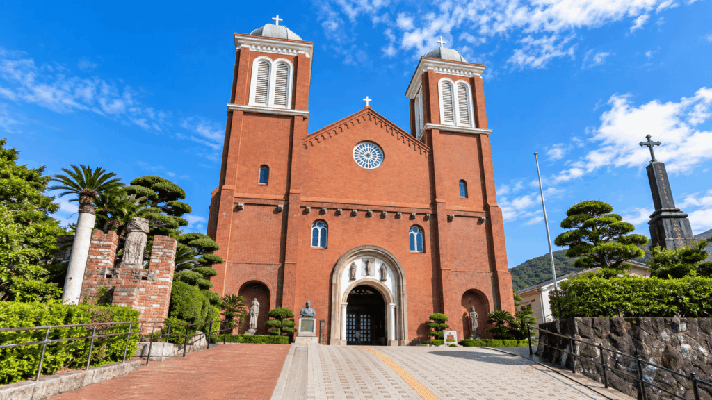 Red-brick facade of Urakami Cathedral in Nagasaki, featuring two bell towers, domed roofs, crosses, and a circular stained-glass window